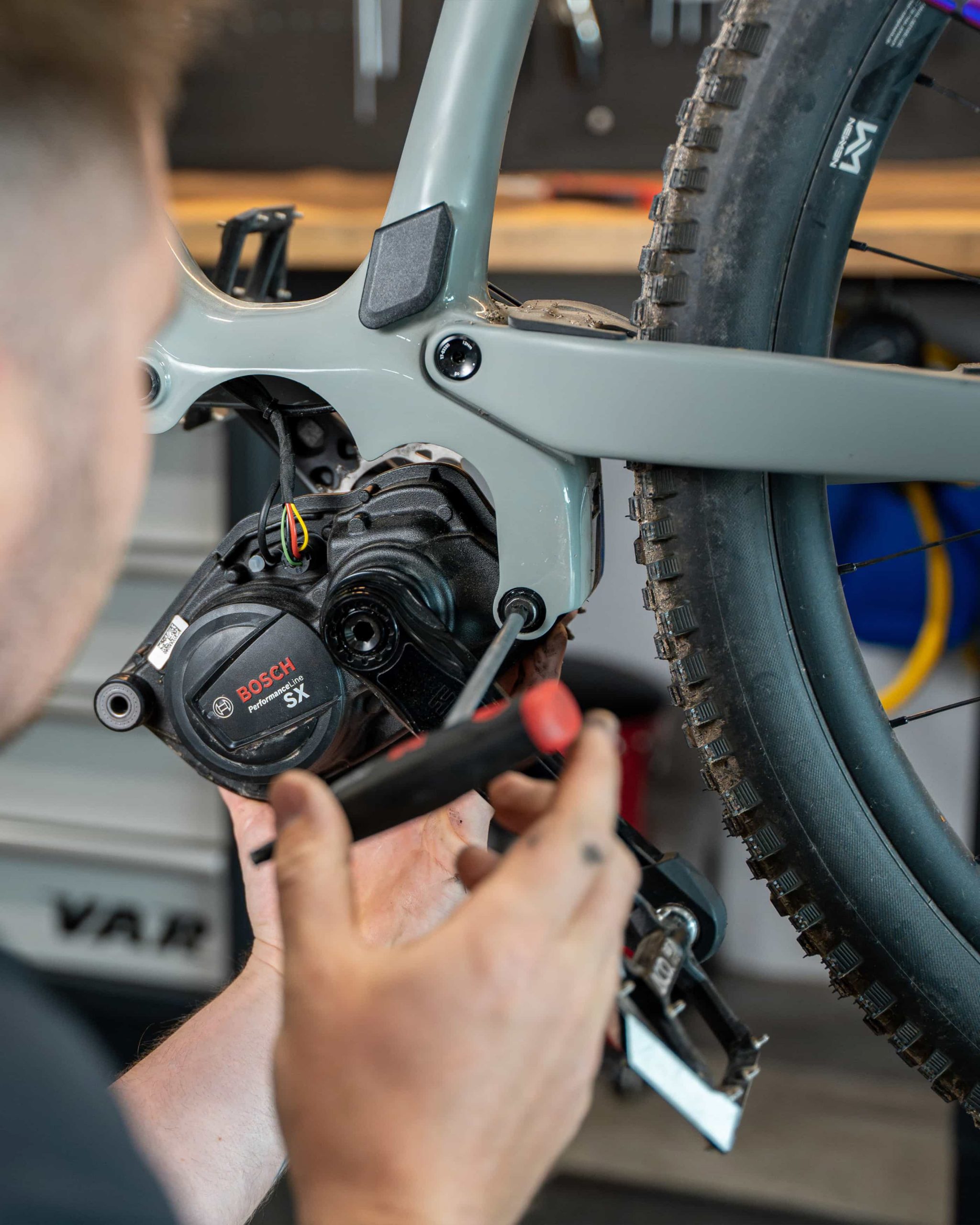 service Nahaufnahme einer Person in einem braunen Kapuzenpullover, die mit einem Schraubenschlüssel die hintere Gangschaltung eines CUBE Fahrrads einstellt. Der Fokus liegt dabei auf der Kette und den mechanischen Teilen des Fahrrads. Perfekt für Mr. Bike-Fans, die gerne selbst Hand anlegen.